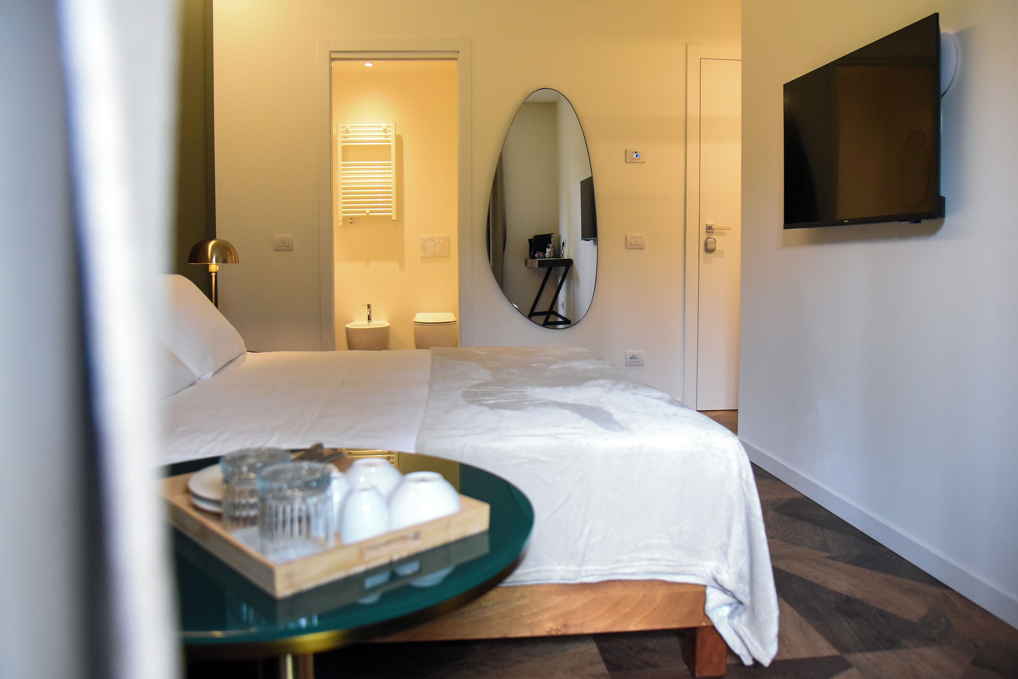 A hotel room with a bed, a bedside table with a tray of glasses, a mirror, a TV mounted on the wall, and a door leading to a bathroom.