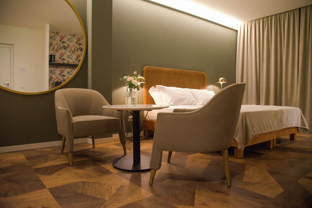 A hotel room with a bed, two chairs, a round table, a mirror, and a nightstand with a vase of flowers.