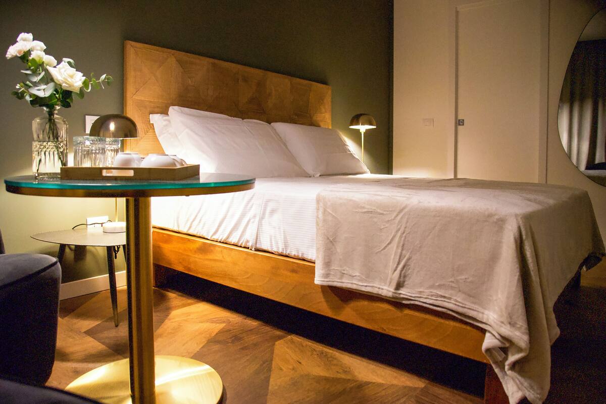A neatly arranged hotel room with a wooden headboard, a round glass table with a tray, a vase with flowers, and a bedside lamp.