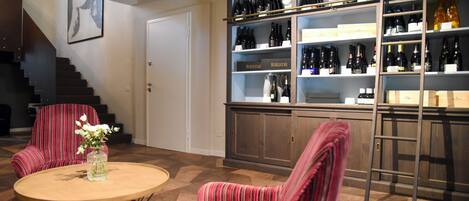 Une pièce avec un plancher en bois, deux chaises à rayures, une table ronde avec un vase, une bibliothèque remplie de bouteilles de vin et un escalier.