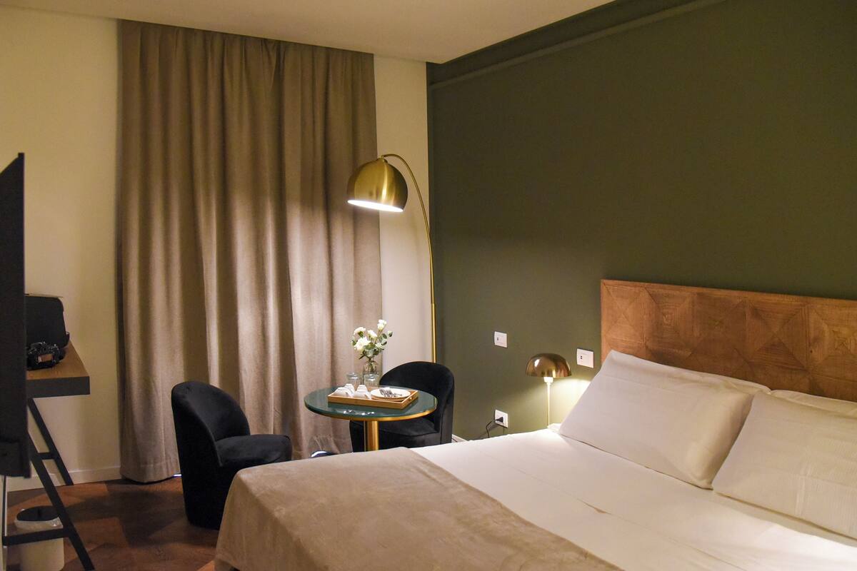 A hotel room with a bed, a desk, a chair, a table with a vase of flowers, and a lamp.