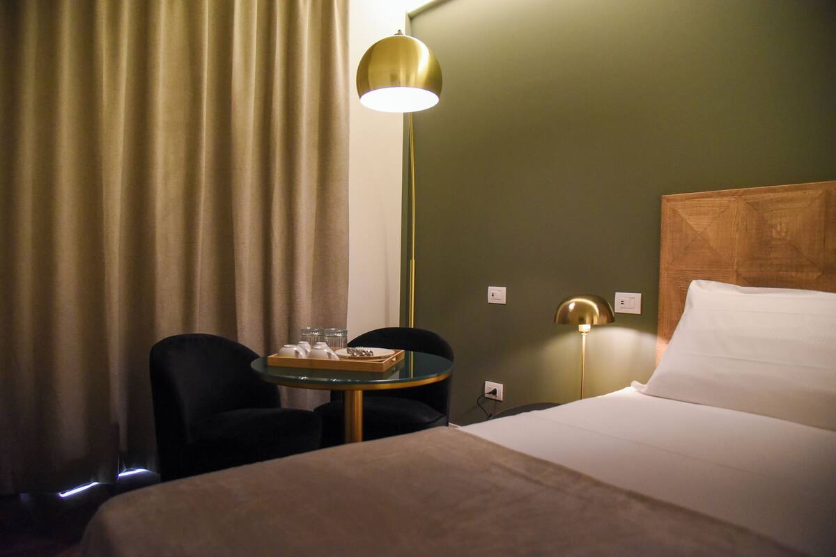 A hotel room with a bed, a bedside table with a tray of items, two chairs, a lamp, and a curtain.