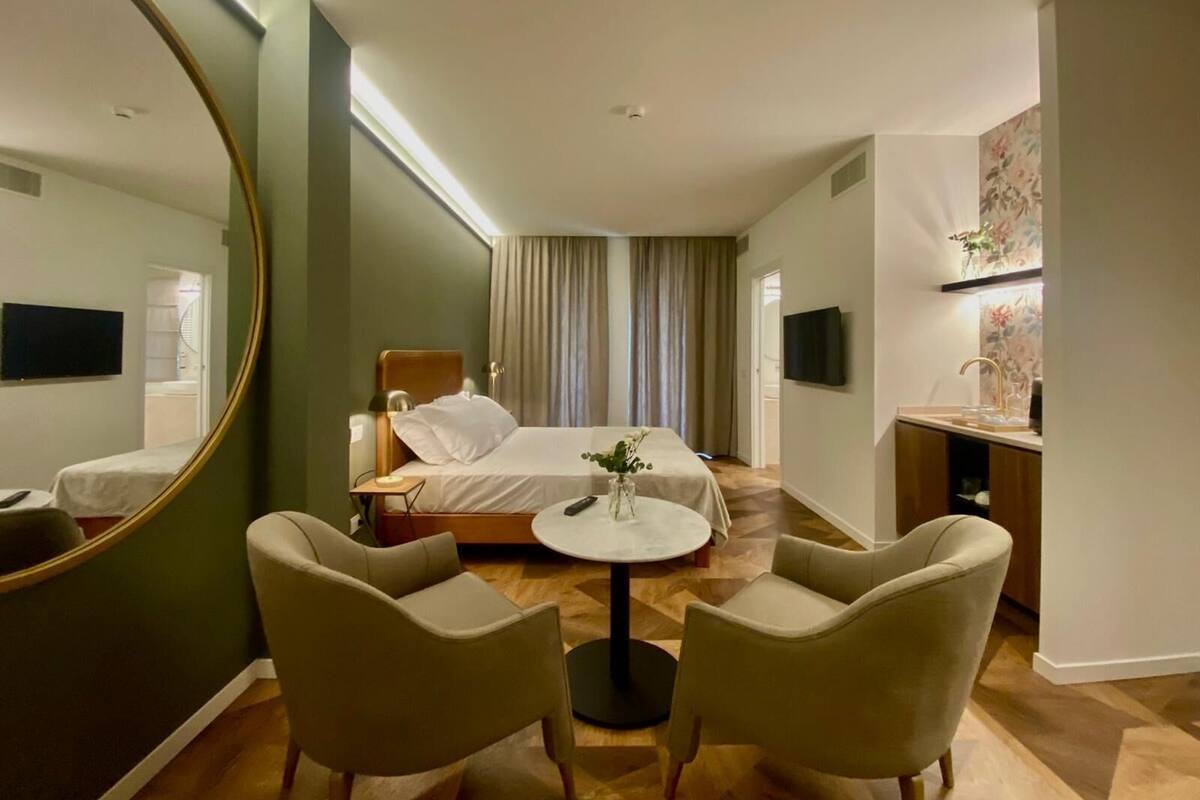 A hotel room with a bed, a round table, two chairs, a large mirror, a TV, and a small desk with a sink.