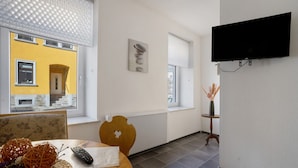 Une pièce comprenant une table à manger, des chaises, un téléviseur fixé au mur, un radiateur et une fenêtre avec des stores.