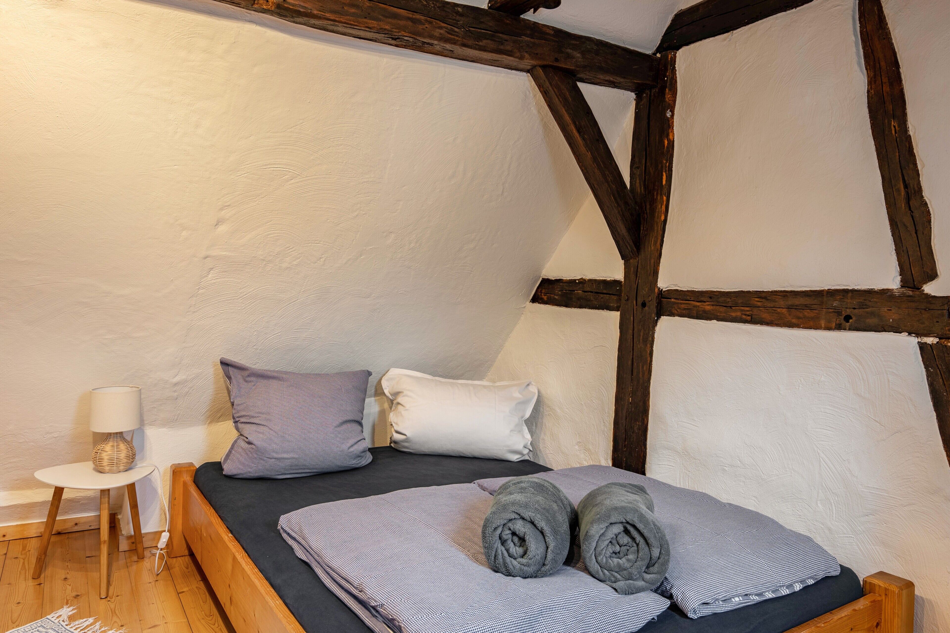 Ein Schlafzimmer mit einem hölzernen Bett, Kissen, einem kleinen Nachttisch mit Lampe und einer Wand mit freiliegenden Balken.