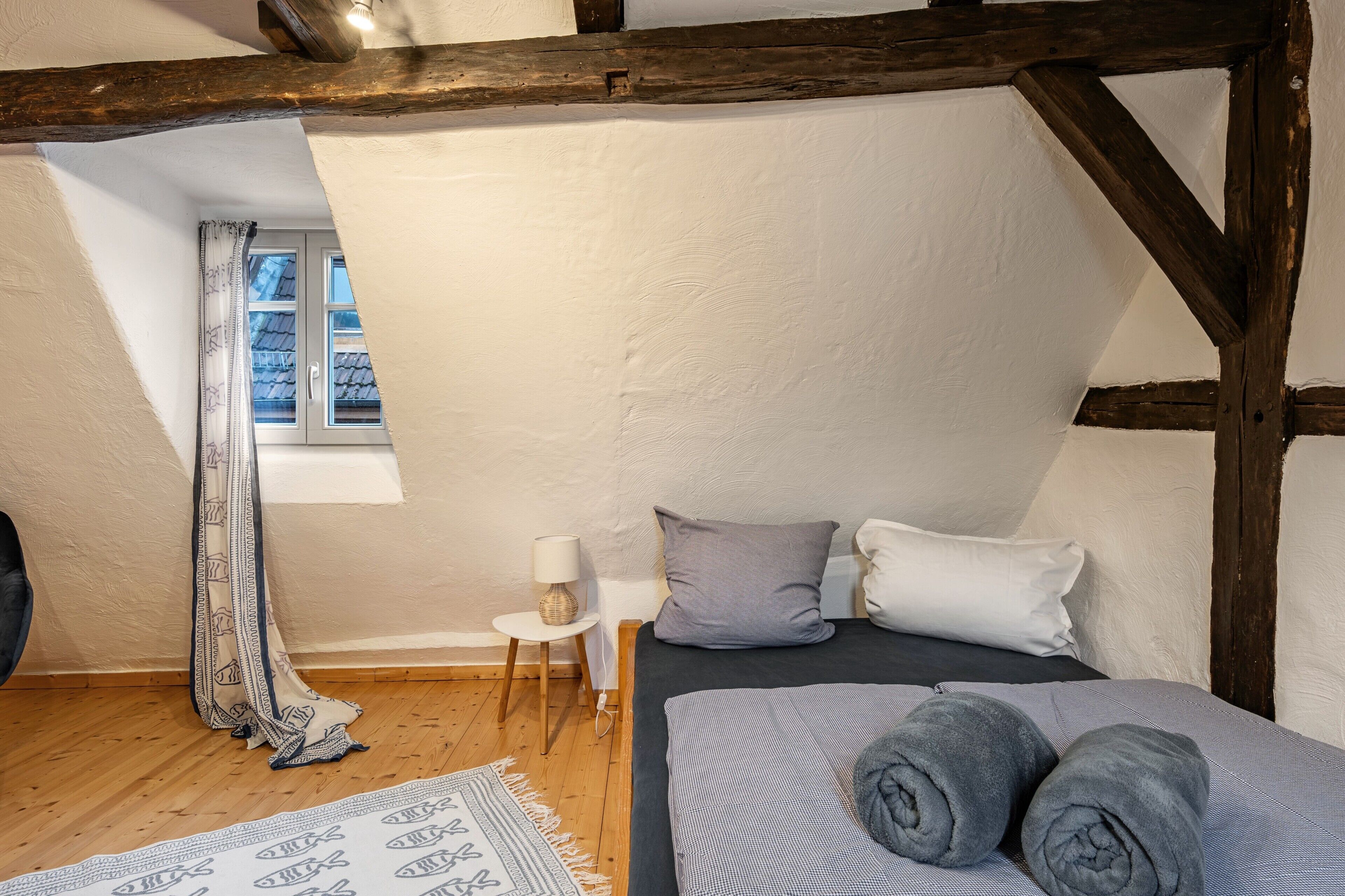 A bedroom with a bed, pillows, a bedside table with a lamp, a window with curtains, and a wooden ceiling beam.