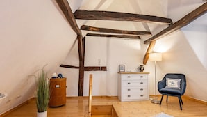 Una habitación con vigas de madera a la vista, un aparador blanco, una silla azul, una planta en maceta y una alfombra estampada.