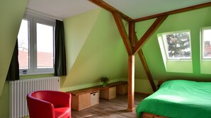 Une chambre avec un plafond en pente, un lit, une chaise rouge, un banc en bois et une fenêtre donnant sur un immeuble.