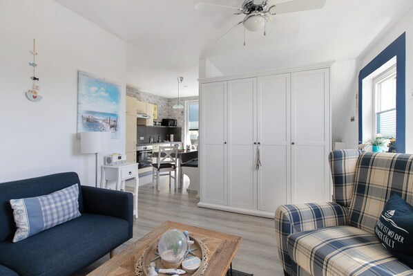A living room with a blue sofa, a plaid sofa, a coffee table with decorative items, a white wardrobe, and a kitchen in the background.