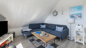 En moderne stue med en hjørnesofa, et træsofabord, et fjernsyn og et hvidt sengebord med en lampe.