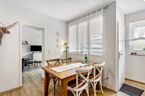 Un coin repas avec une table en bois, des chaises blanches et une petite table d’appoint sur laquelle se trouve une bouteille.