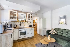Une cuisine compacte avec des meubles blancs, un four encastré et un réfrigérateur. On y trouve un canapé vert agrémenté d’un coussin à motifs et une petite table ronde sur laquelle est posée une bougie.