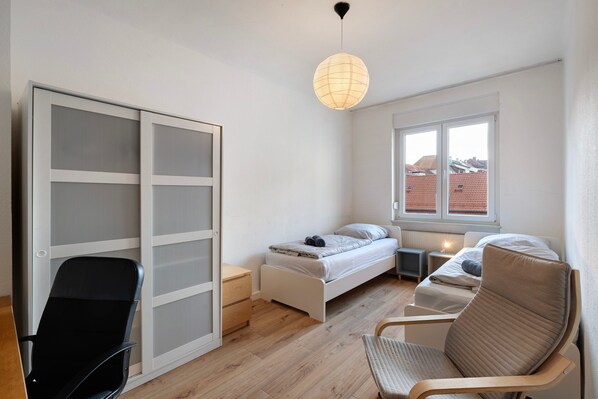 Une chambre à coucher comprenant un lit, deux tables de chevet, une chaise, une armoire et une fenêtre donnant sur des immeubles.