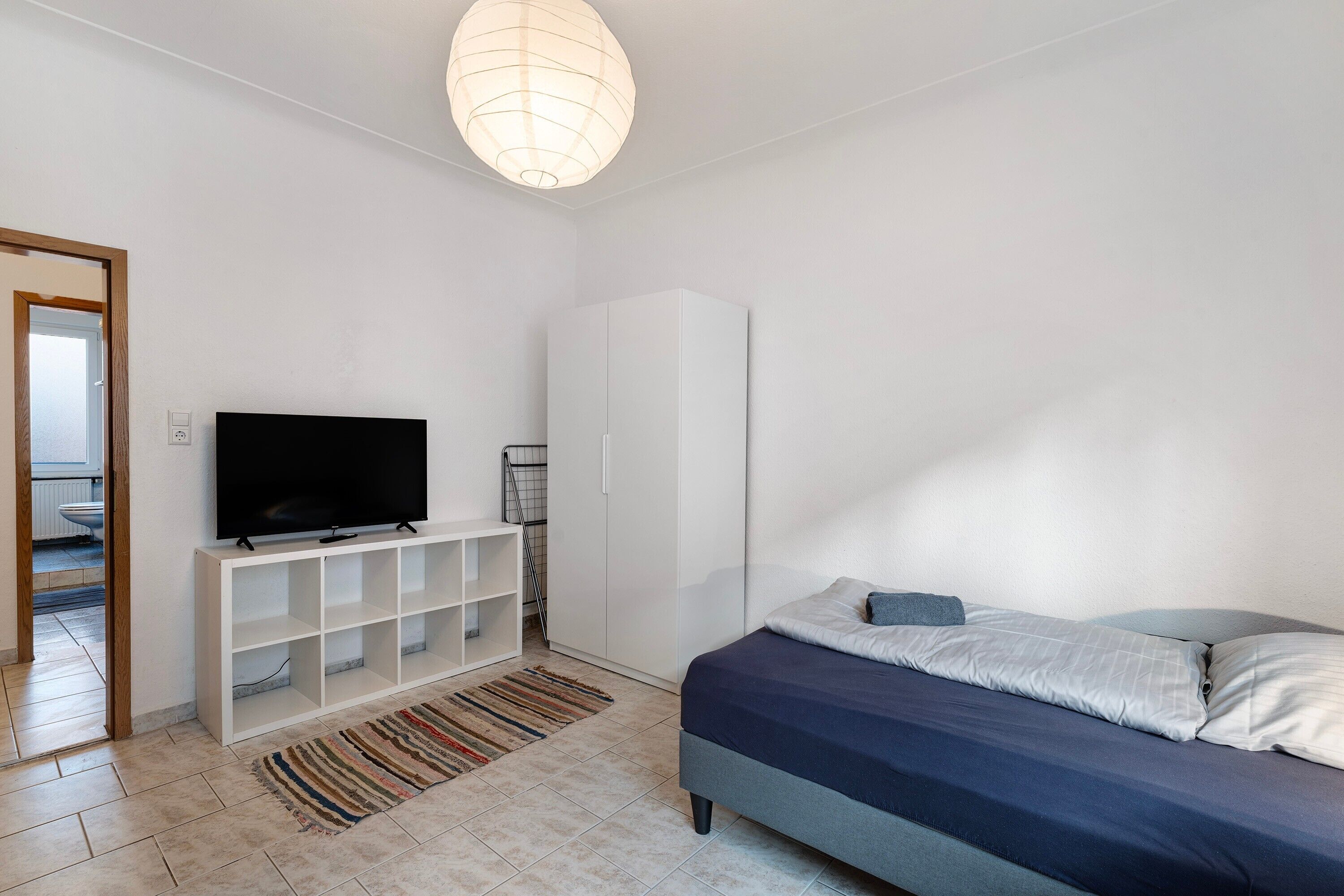A bedroom with a bed, a TV on a white stand, a wardrobe, and a door leading to a bathroom.