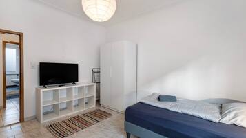 A bedroom with a bed, a TV on a white stand, a wardrobe, and a door leading to a bathroom.