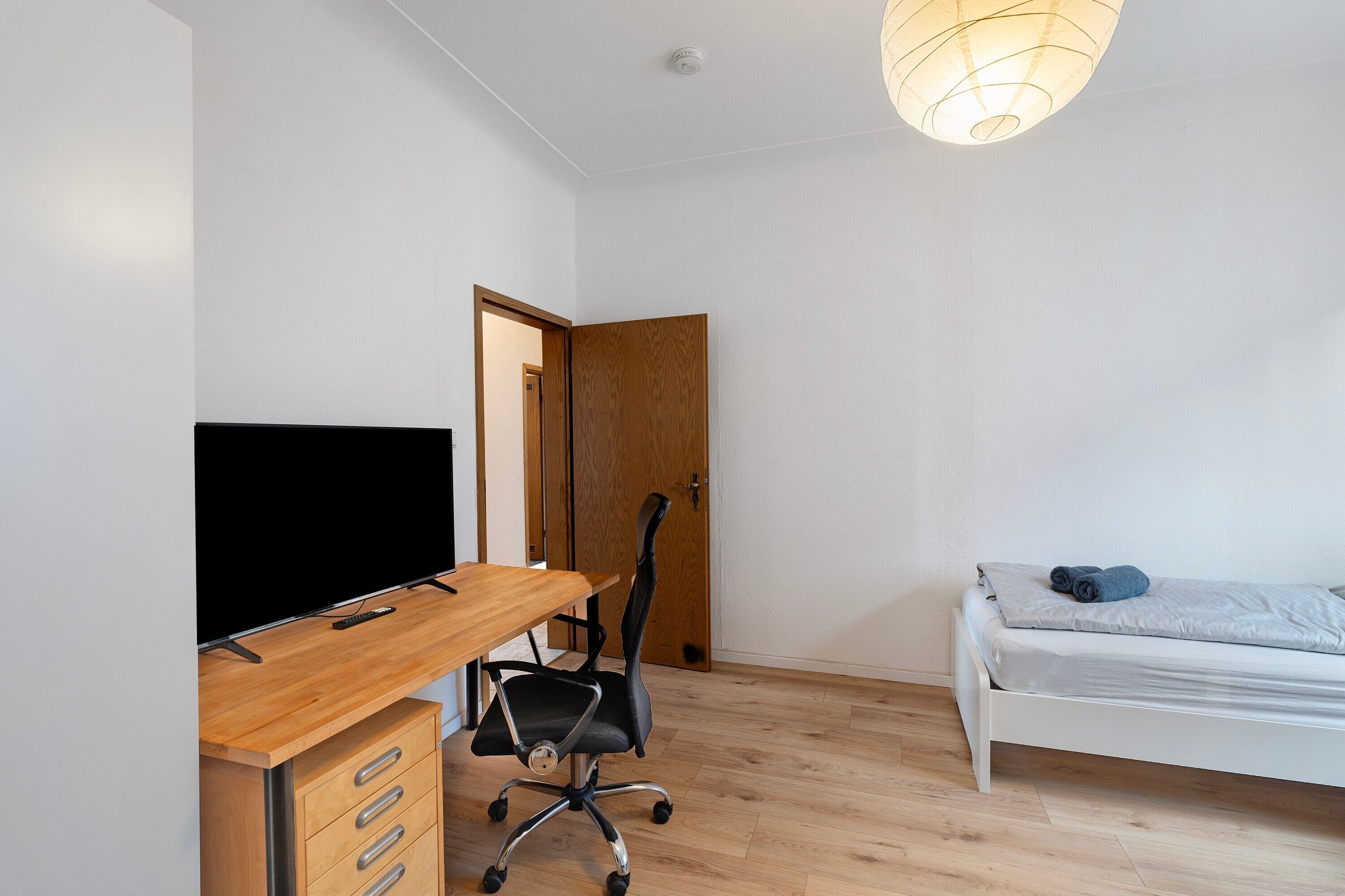 A modern room with a wooden desk, a black office chair, a white bed, and a wooden door.