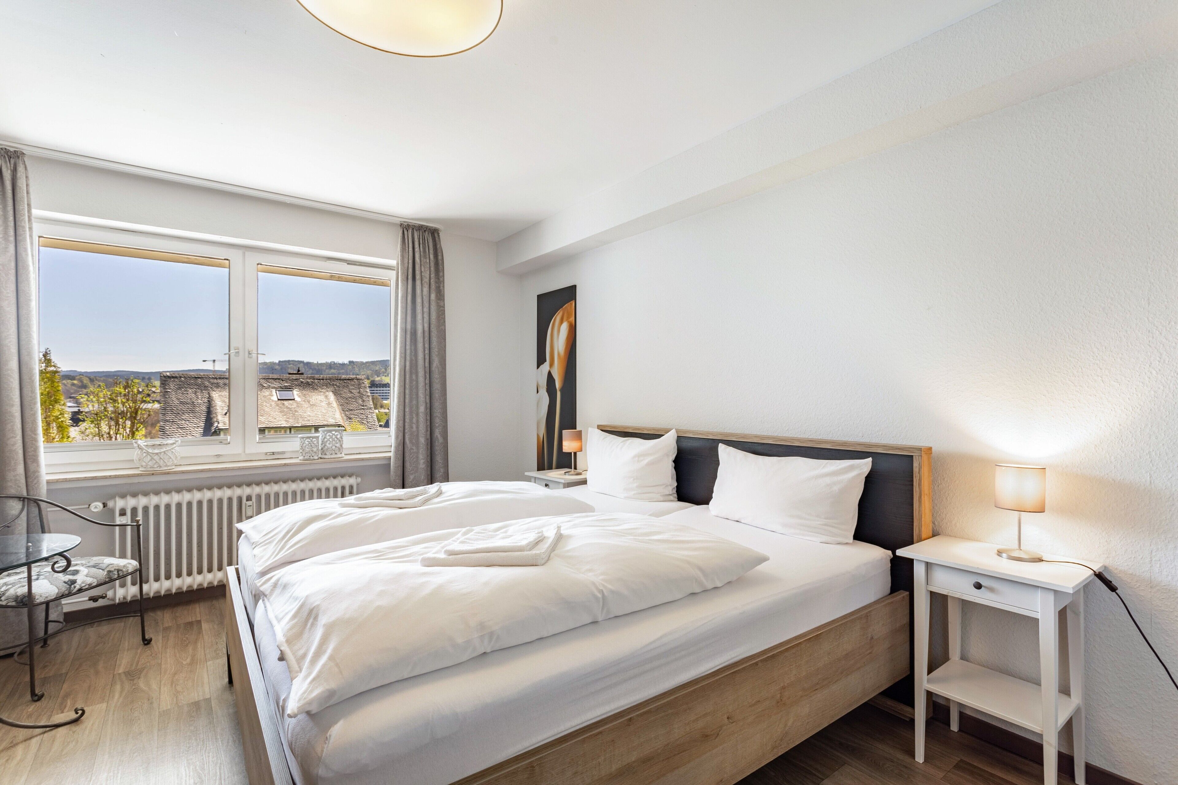 Ein Schlafzimmer mit einem großen Bett, einem Nachttisch mit Lampe, einem Sessel und Blick auf die Stadt durch ein Fenster.
