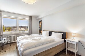 Um quarto com uma cama grande, uma mesa de cabeceira com abajur, uma cadeira e vista para a cidade através de uma janela.