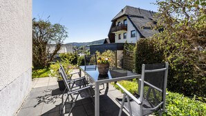 A patio with a glass table and chairs, a potted plant, and a view of a house and hills. - Holiday Apartments Löwenstein FeWo 1 with Private Terrace & Garden and Wi-Fi (Willingen (Upland))