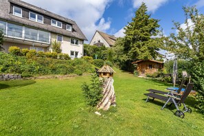 Uma área residencial com gramado bem cuidado, um comedouro de madeira para pássaros e um par de espreguiçadeiras.