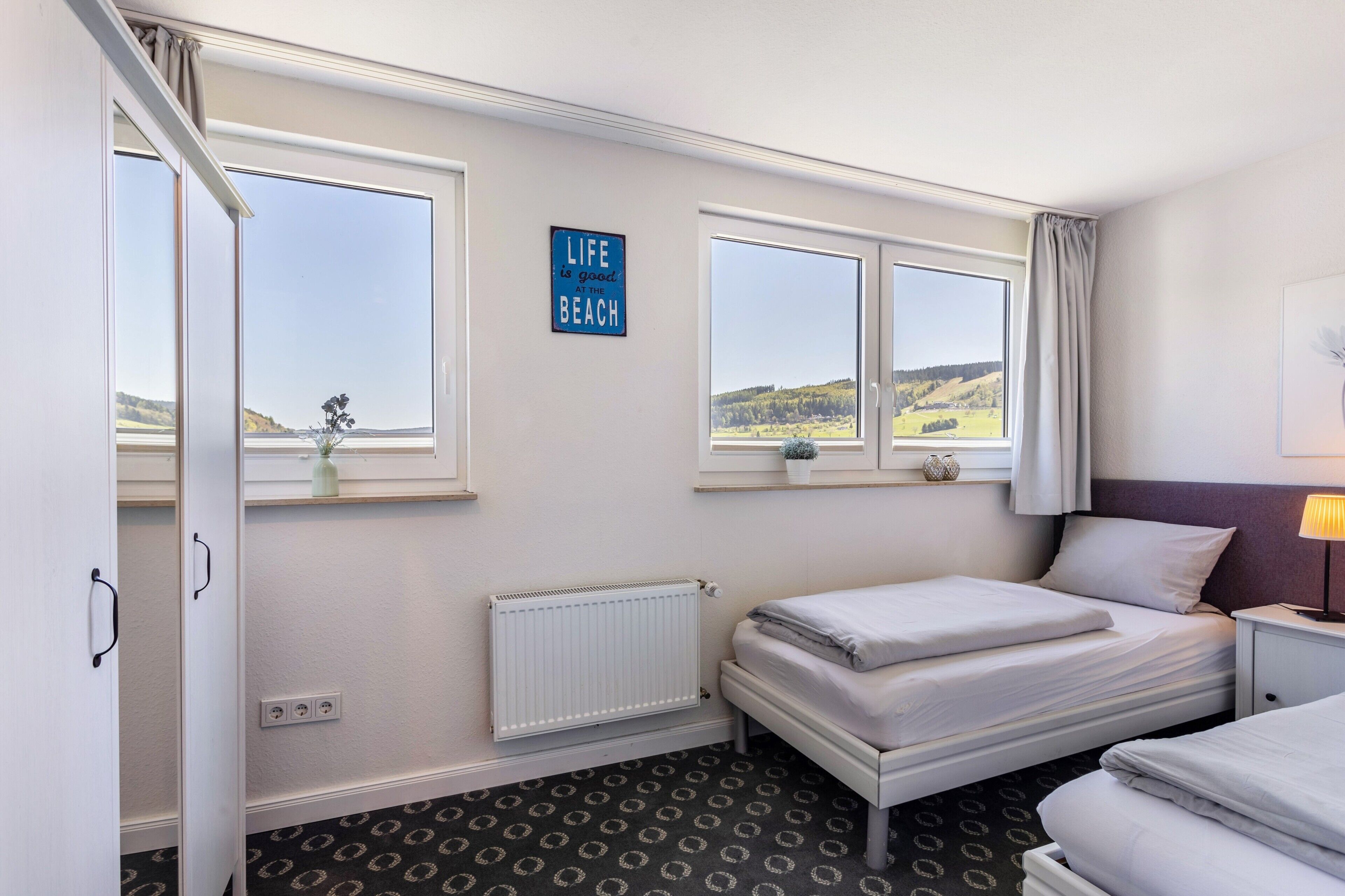 Een hotelkamer met twee bedden, een radiator, een raam met uitzicht op het landschap, en een bordje met de tekst 'het leven is goed'.