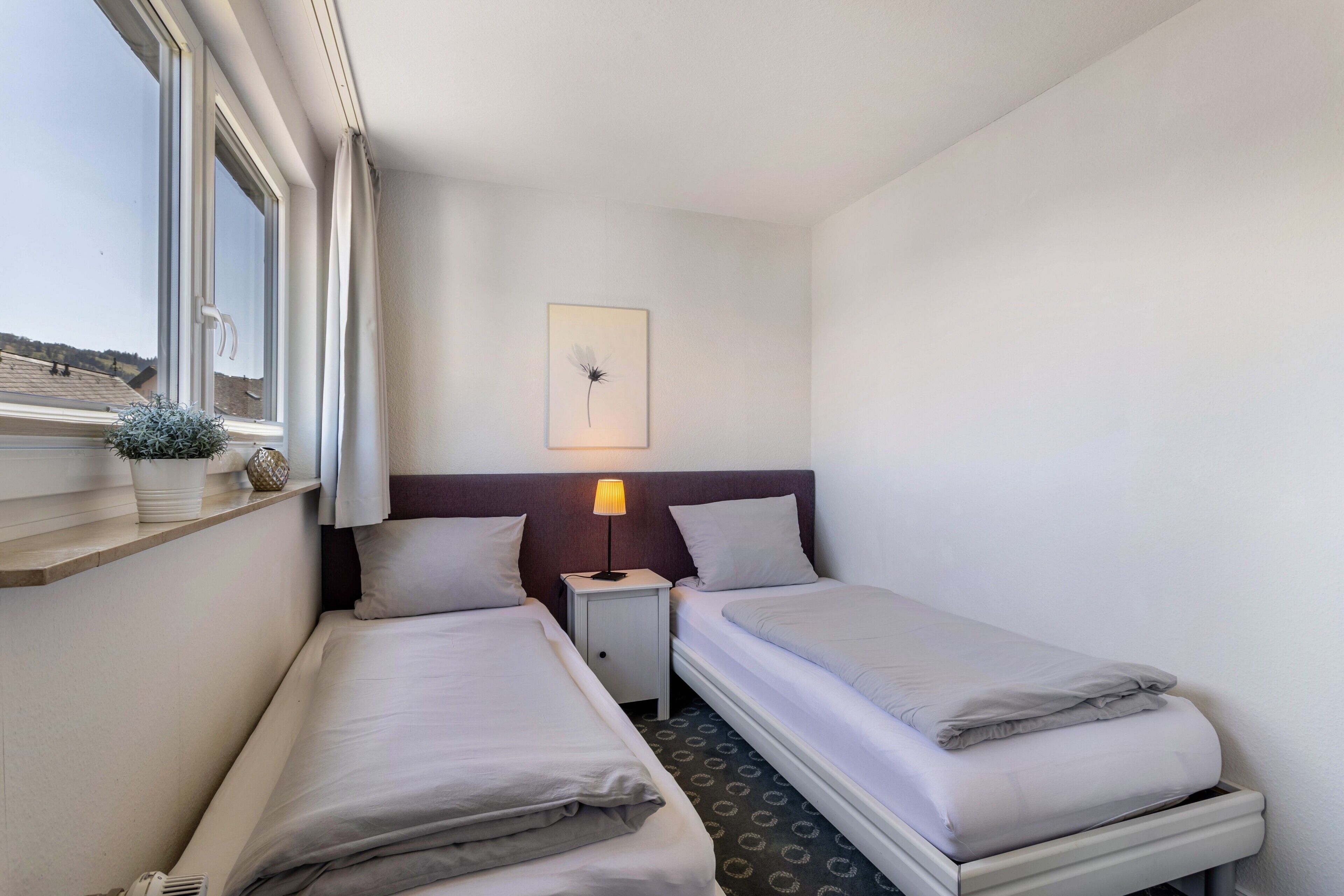 Une chambre d’hôtel avec deux lits simples, une table de chevet avec une lampe, un tableau encadré au mur et une fenêtre donnant sur l’extérieur.