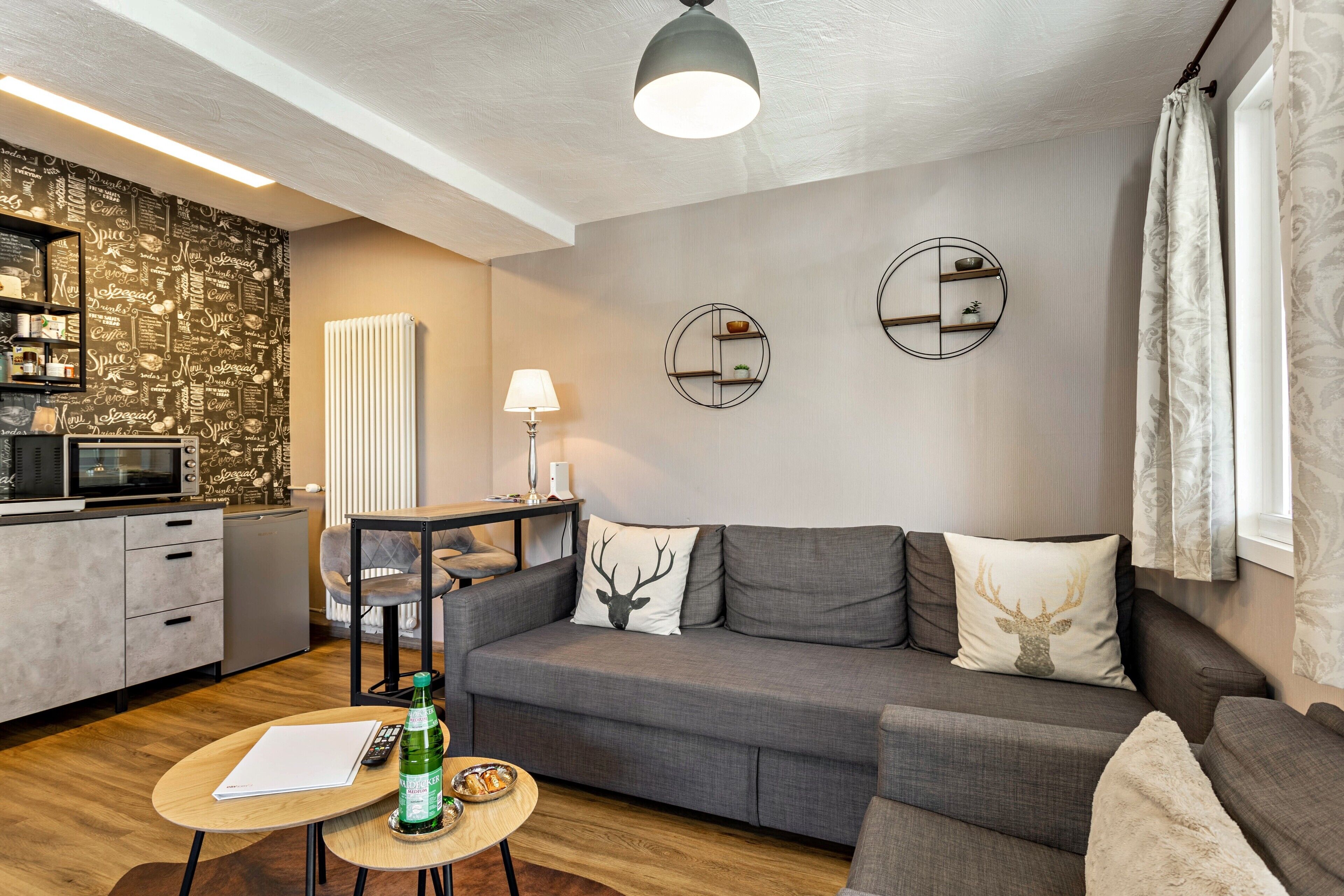 A modern living room with a grey sofa, a small table, a lamp, and a kitchen area with a microwave and cabinets.