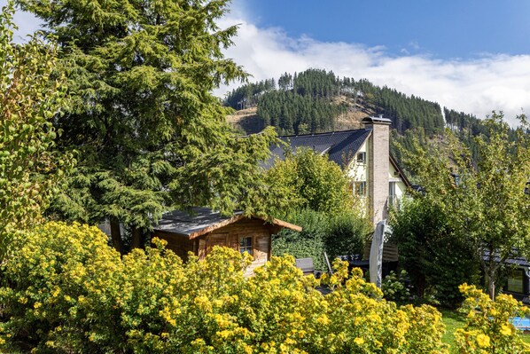 A cabin with a sloped roof, surrounded by dense green trees and a mountain backdrop.