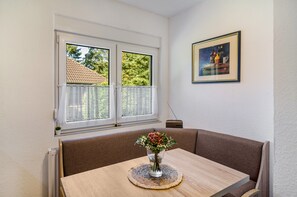 Une pièce comprenant une table, un vase avec des fleurs, un tableau encadré au mur et une fenêtre donnant sur des arbres et un bâtiment.