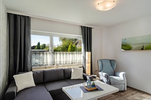 Un salon moderne avec un canapé gris, une table basse blanche, un fauteuil bleu et une grande fenêtre avec des rideaux.