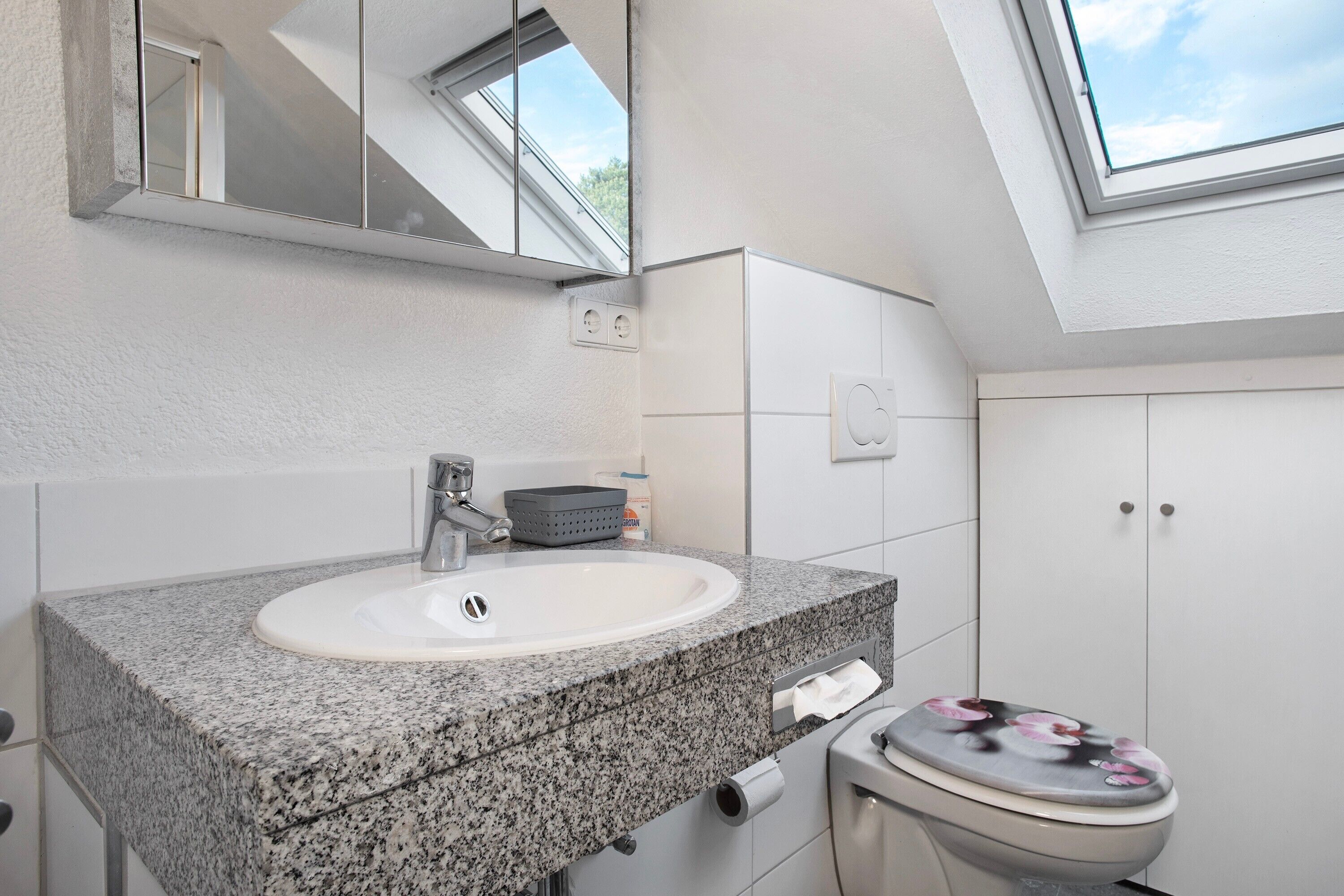 Un baño con lavamanos de encimera de granito, un espejo, una ventana y un inodoro con un asiento decorado con motivos florales.