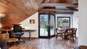 A room with a wooden ceiling, a dining area with chairs and a table, a desk with a chair, and a large window with a view.