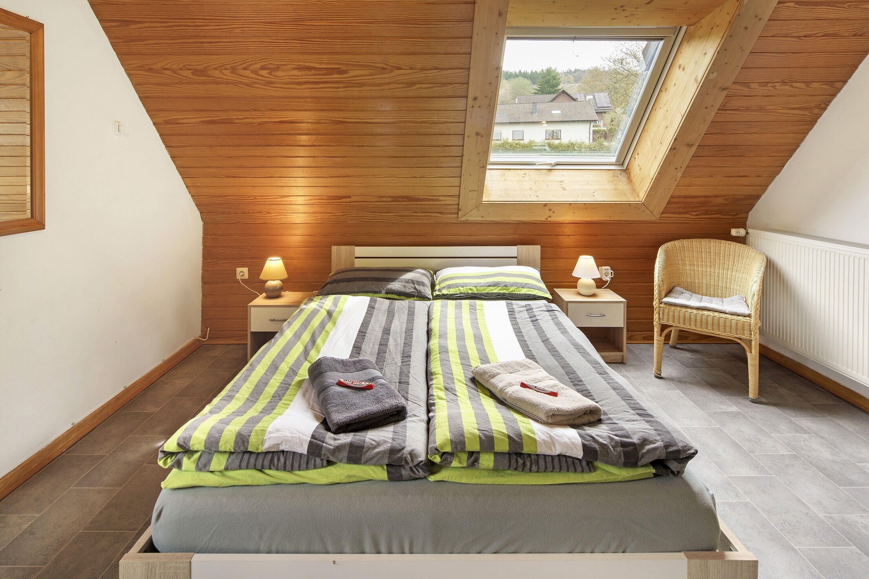 Ein Schlafzimmer mit schrägem Dach, einem Oberlicht, einem Bett mit gestreifter Bettwäsche, zwei Nachttischen mit Lampen, einem Korbstuhl und einer Heizung.