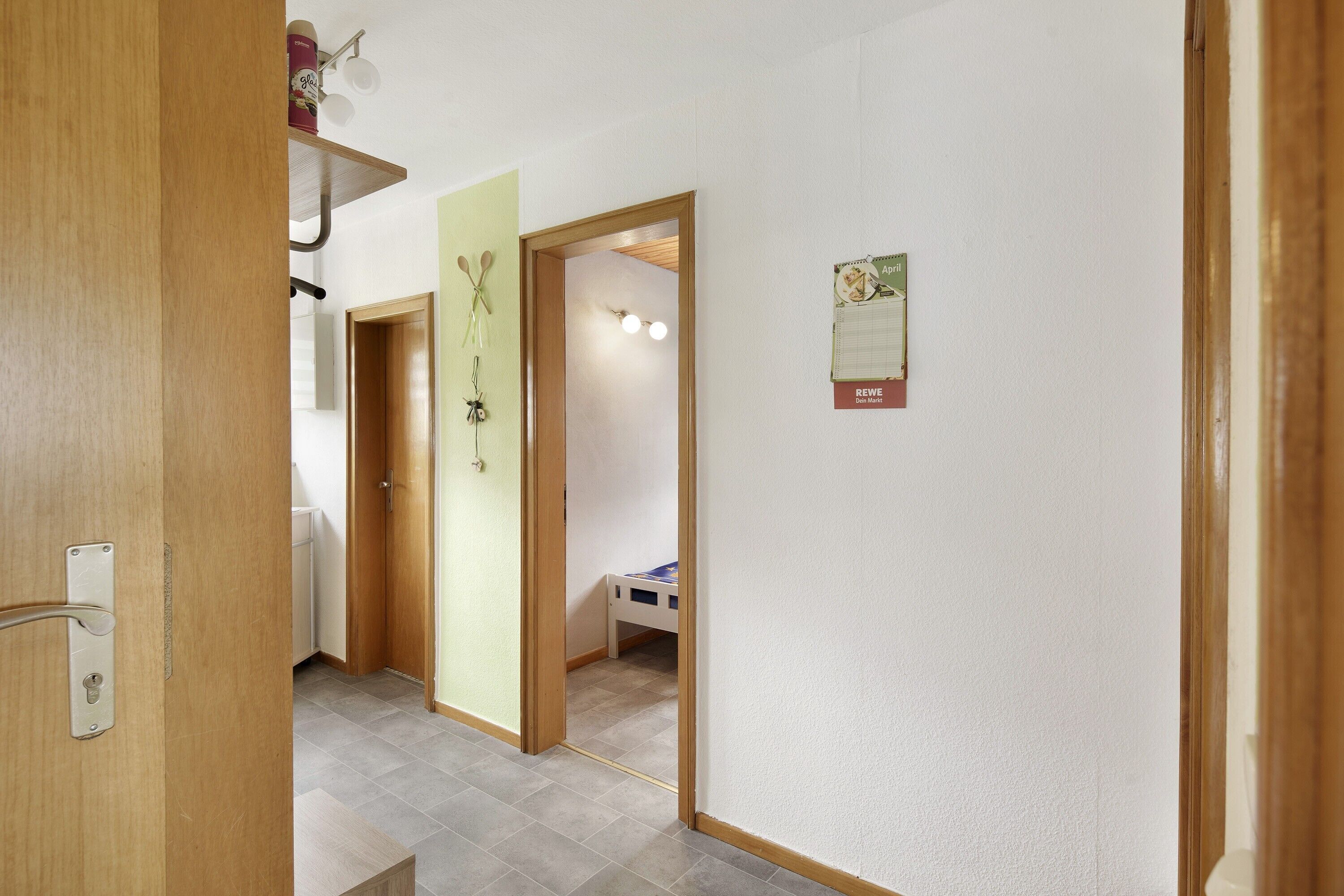 Un pasillo con una puerta de madera, una cama pequeña y un calendario en la pared.