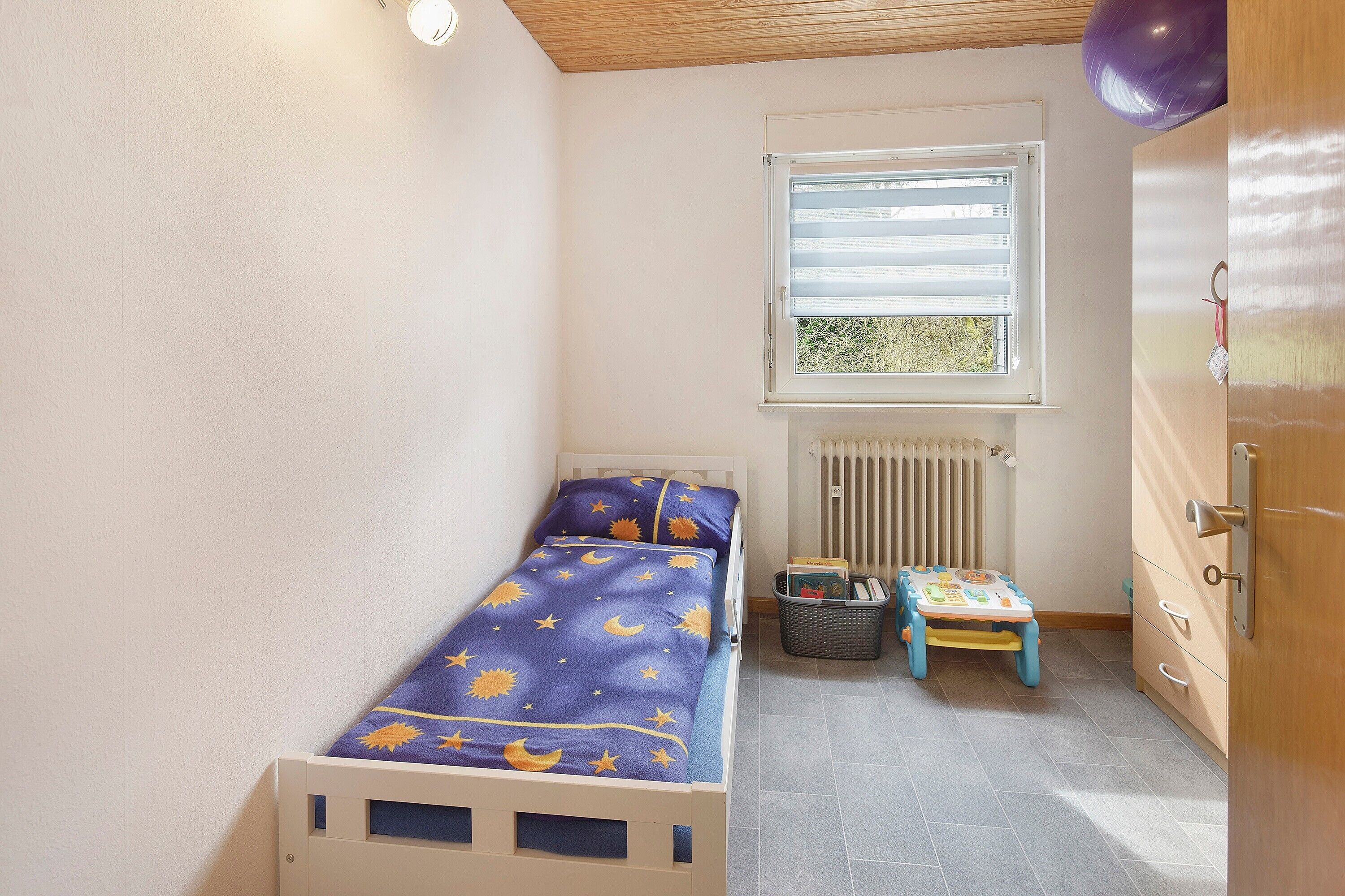 Ein kleines Zimmer mit einem Einzelbett, einem Fenster, einer Heizung und einem kleinen Tisch.