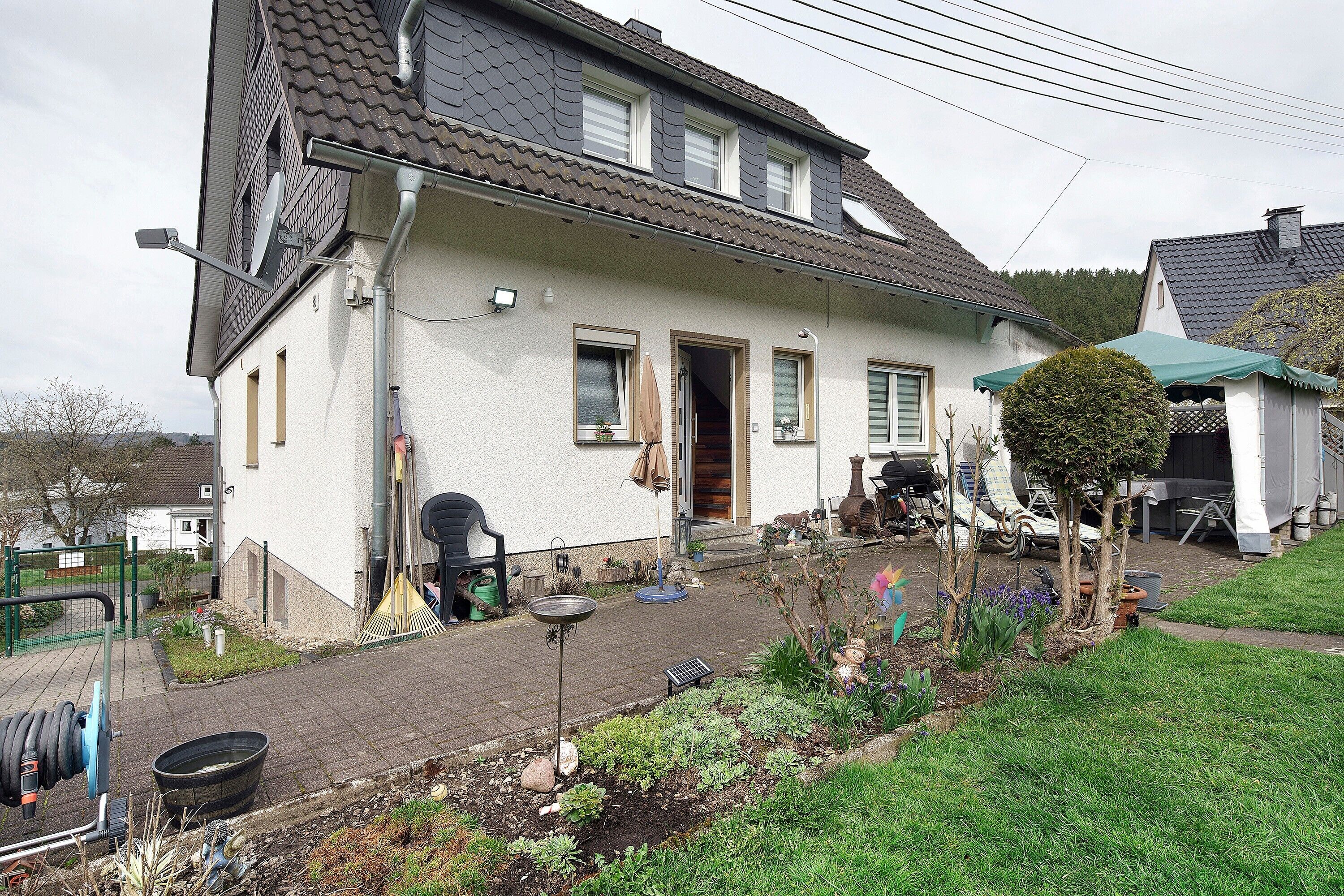 Une maison à deux étages avec un toit en pente, une porte d’entrée et un petit jardin agrémenté de plantes et d’un abreuvoir pour oiseaux.
