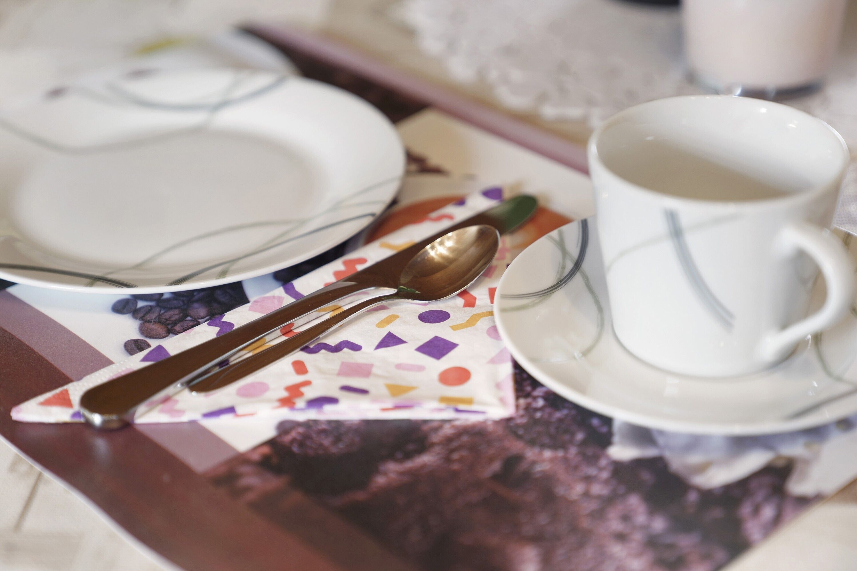 Ein Tisch mit einem weißen Teller, einer weißen Tasse und einem Löffel auf einer Serviette mit farbigem geometrischen Muster.
