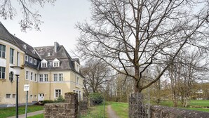 Un chemin menant à un bâtiment doté d’un mur en pierre et d’une grille en métal, entouré d’arbres et d’une zone herbeuse.