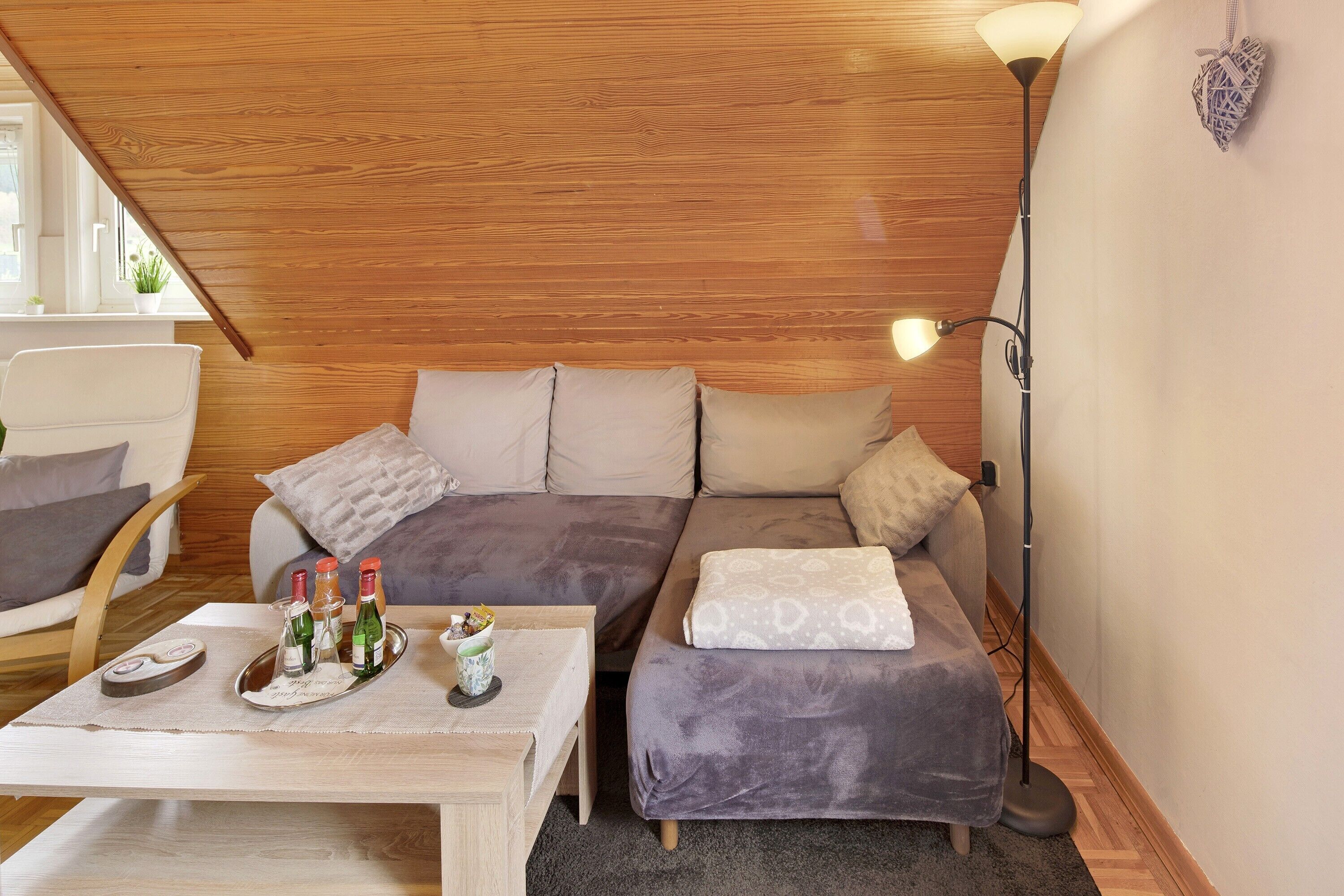 Ein gemütliches Zimmer mit Holzbalkendecke, einer Couch, einem Sessel, einem Tisch mit Flaschen und einer Stehlampe.