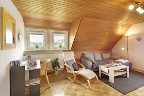A cozy loft-style room with a wooden ceiling, a sofa, a rocking chair, a small table, and a window with a view.