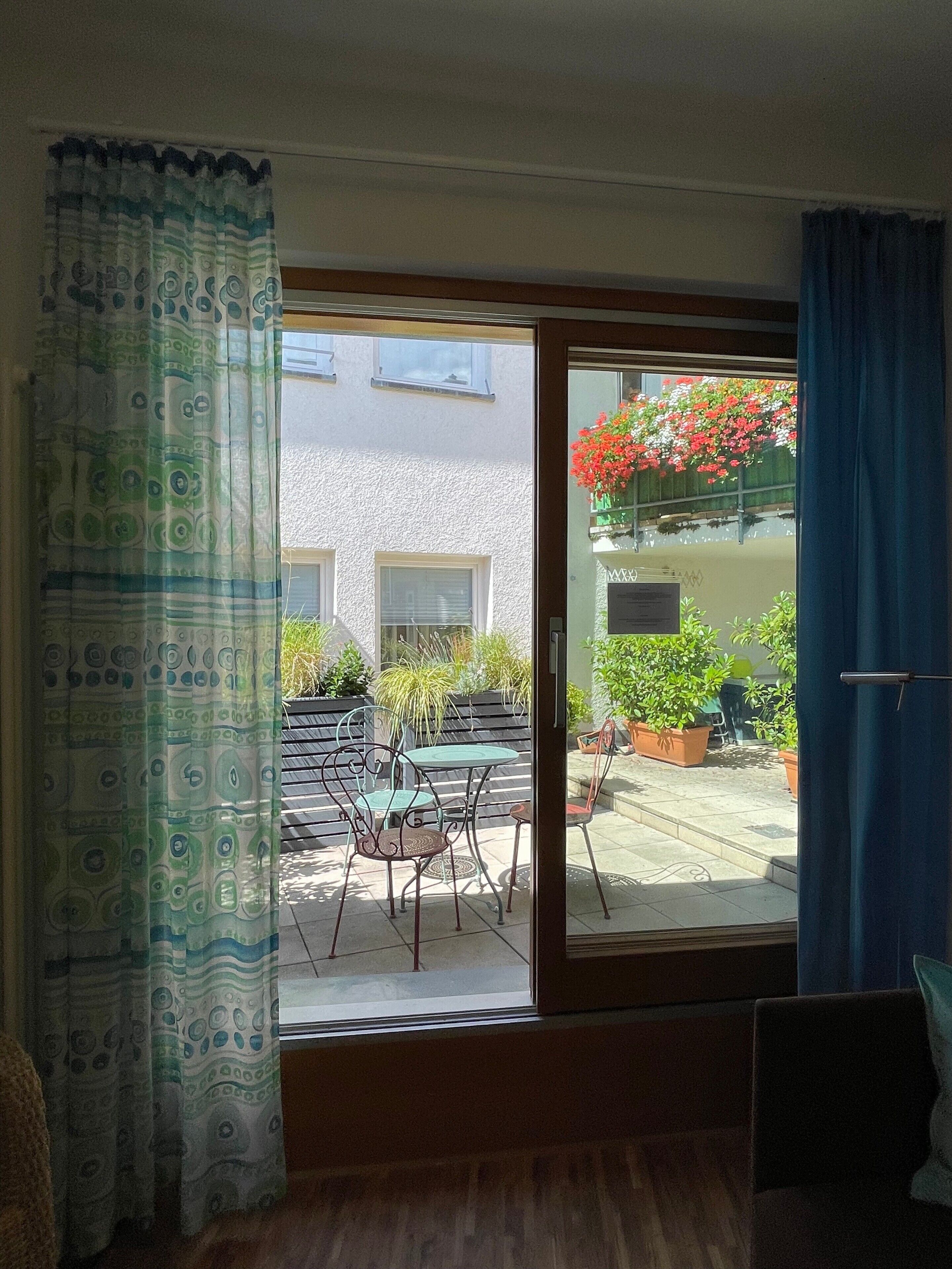 Una stanza con una porta vetrata scorrevole che dà su un patio esterno con tavolo e sedie, piante in vaso e vista su un edificio con finestre.