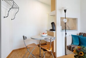 Ett kompakt kök med ett runt bord, två stolar, en skål med frukt, en vattenkokare och en kaffemaskin.