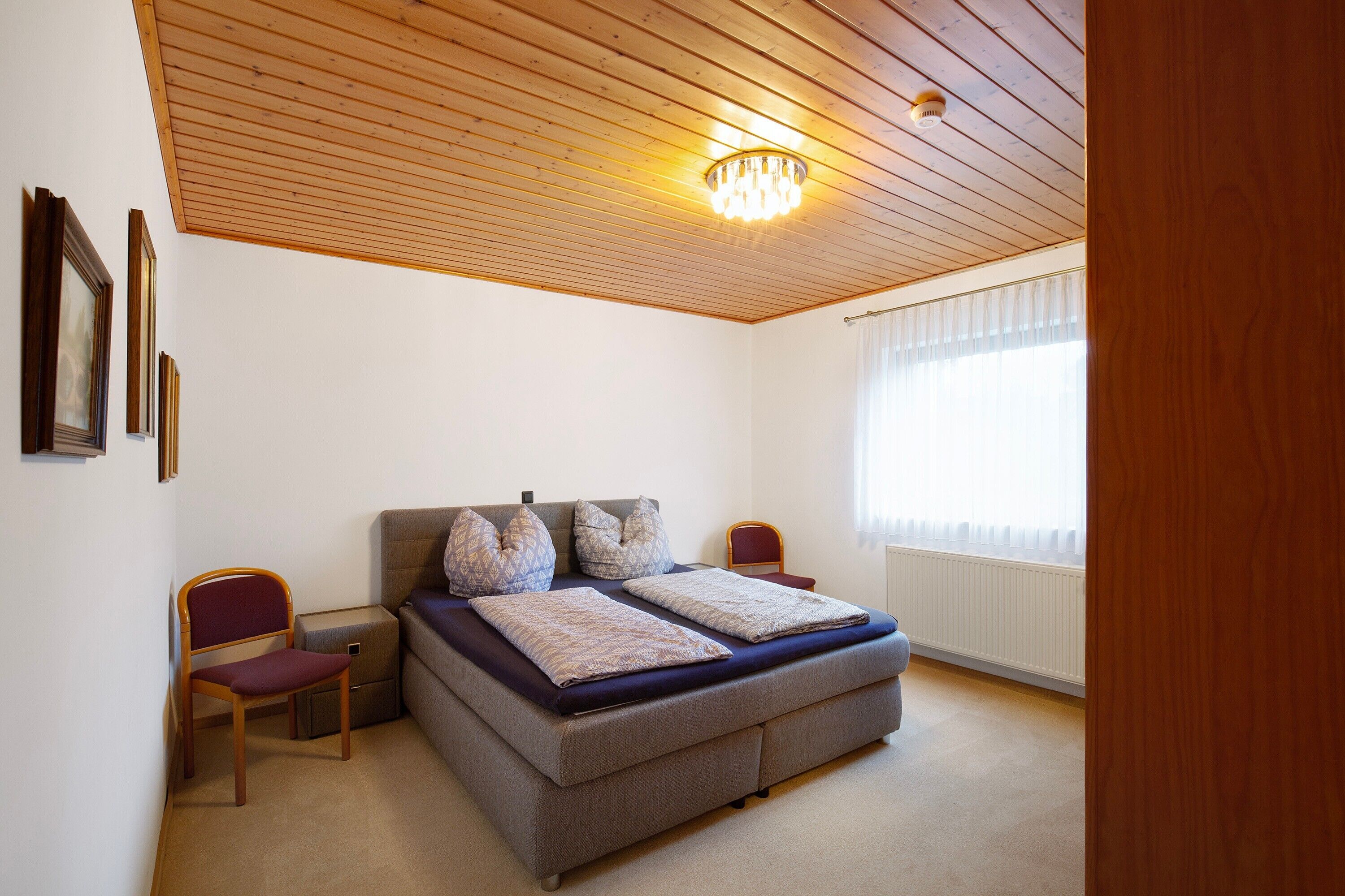 A bedroom with a bed, two chairs, a wooden ceiling, and a window with curtains.