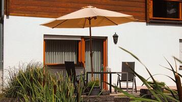 Une terrasse avec une table et des chaises, un parasol et une fenêtre avec des rideaux.