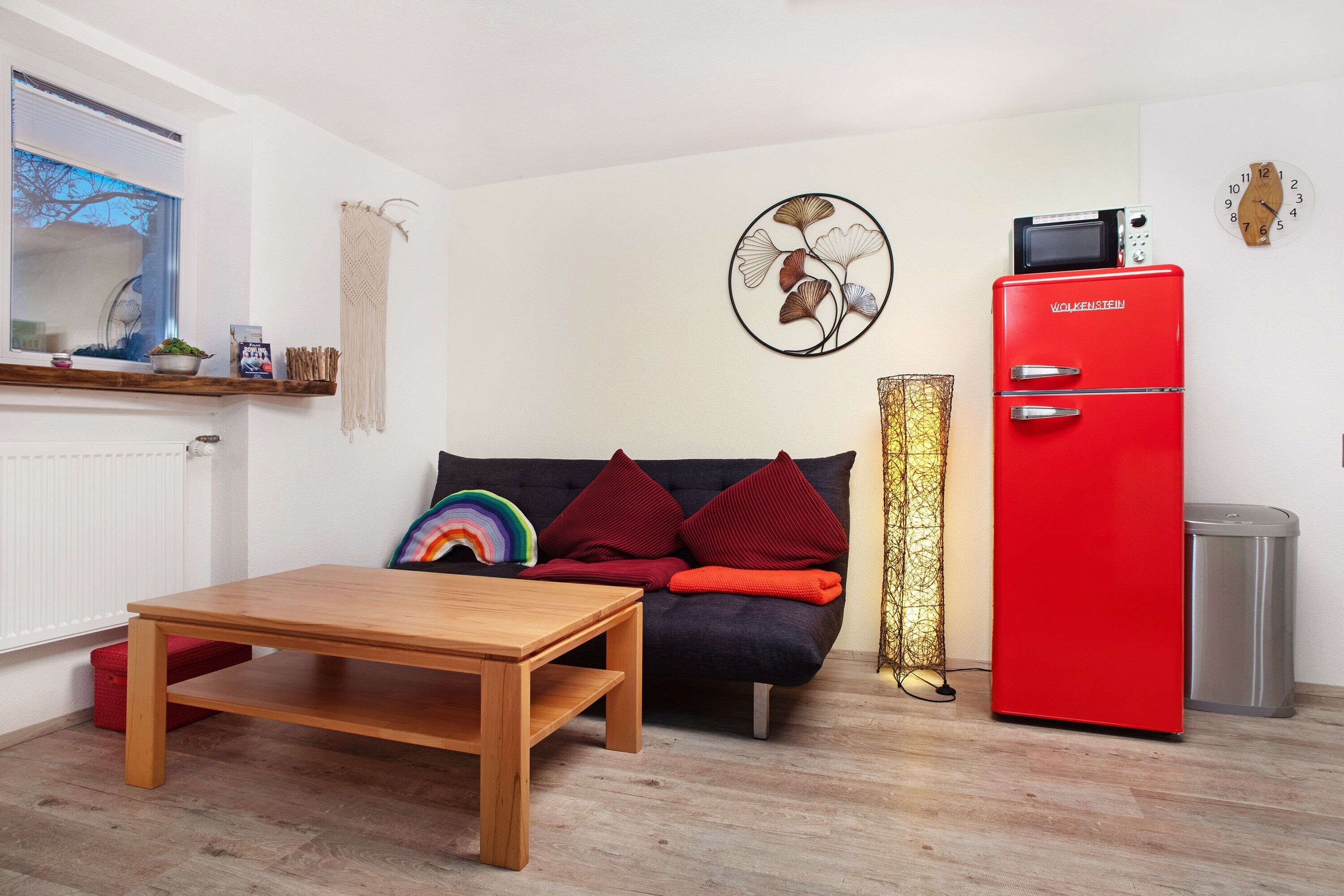 A small living space with a wooden table, a sofa with colorful pillows, a red refrigerator, and a wall clock.