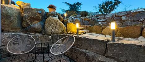 A stone patio with two wicker chairs and a table, surrounded by large rocks and a potted plant.