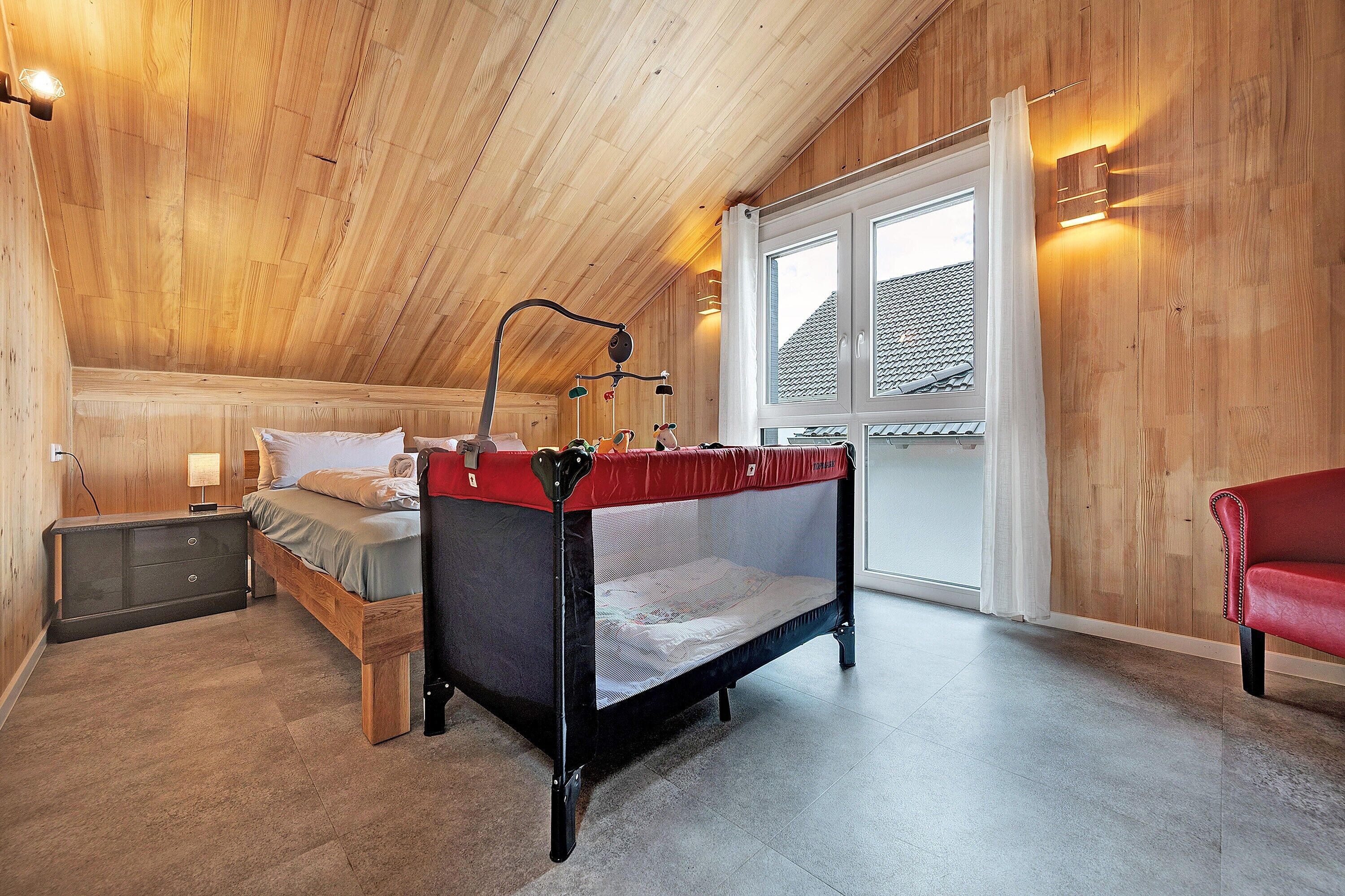 Ein Schlafzimmer mit Holzbalkendecke, einem Himmelsbett, einem Nachttisch, einem Fenster mit Vorhängen und einem roten Sessel.