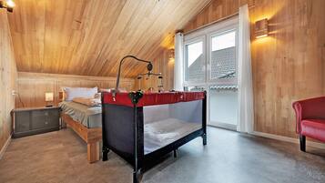 Ein Schlafzimmer mit Holzbalkendecke, einem Himmelsbett, einem Nachttisch, einem Fenster mit Vorhängen und einem roten Sessel.