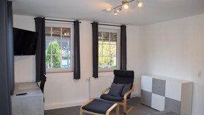 Ein Zimmer mit einem einzelnen schwarzen Sessel, einem kleinen Tisch, einem an der Wand montierten Fernseher und einem fenster mit Vorhängen.