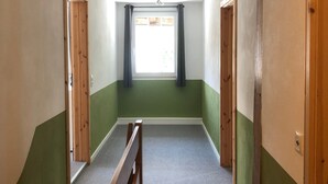 Ein schmaler Flur mit einer hölzernen Bank, einem Fenster mit Vorhängen und einer Tür auf der rechten Seite.